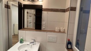 Une salle de bain avec un lavabo, un miroir et des toilettes, tous de couleur blanche. On trouve une bouteille de savon liquide sur le lavabo et une bouteille de gel hydroalcoolique sur le rebord de la fenêtre.