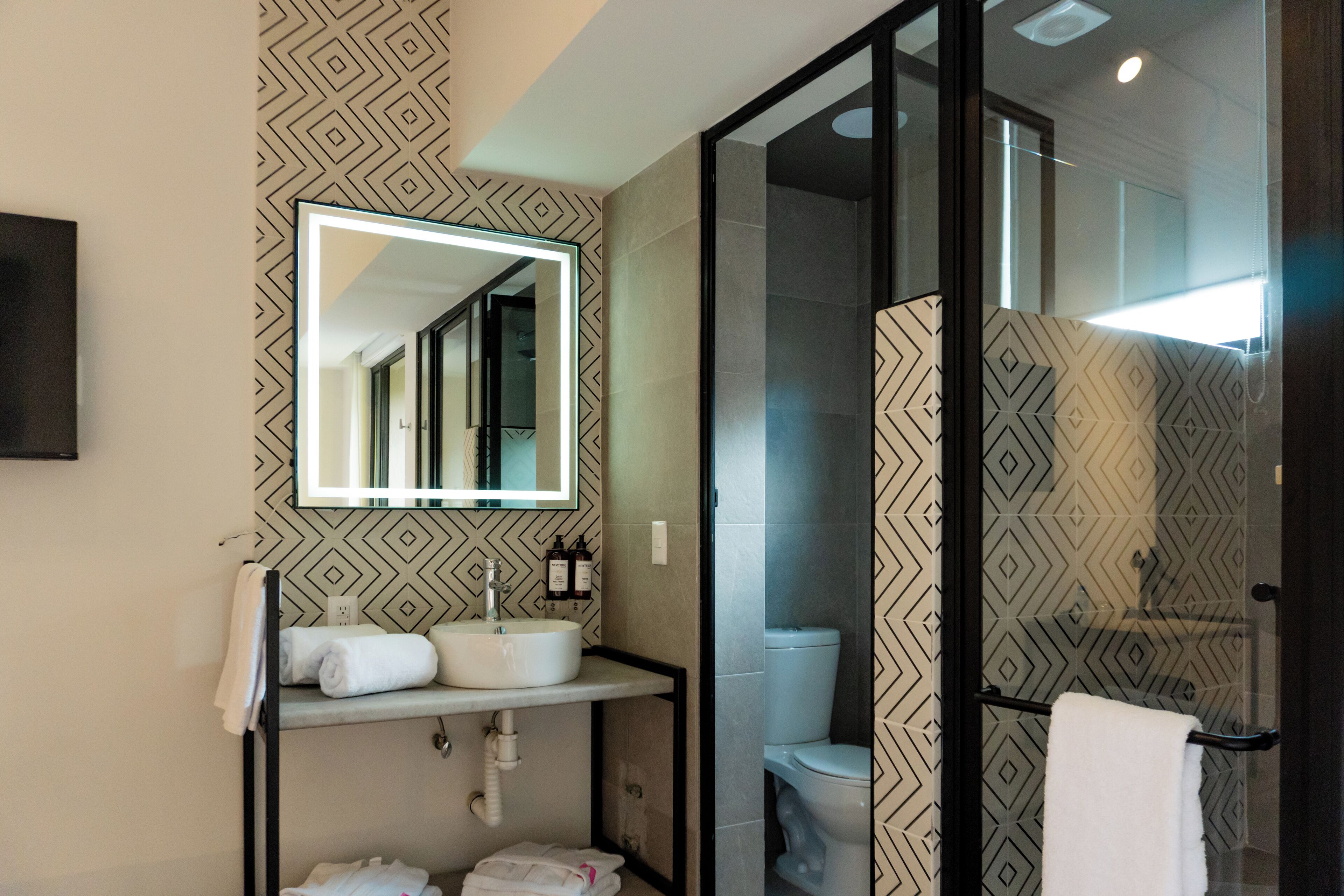 Une salle de bain moderne avec un mur orné d’un motif géométrique, un grand miroir, un lavabo avec un porte-serviettes et une douche à l’italienne.