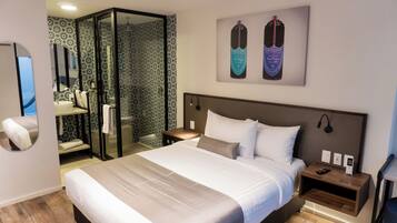 Una habitación de hotel moderna con una cama grande, cabina de ducha de vidrio, baño con espejo y dos objetos enmarcados en la pared.