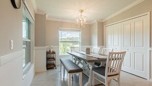 A dining area with a large table, chairs, a bench, a chandelier, and a view through a window.