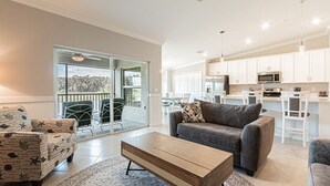 A modern living room with a sofa, armchair, and coffee table. There is a sliding glass door leading to a balcony with outdoor furniture.