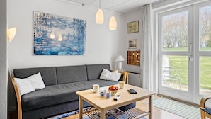 Ein modernes Wohnzimmer mit einem grauen Sofa, einem Holztisch und einem großen Fenster mit Blick auf einen Garten.
