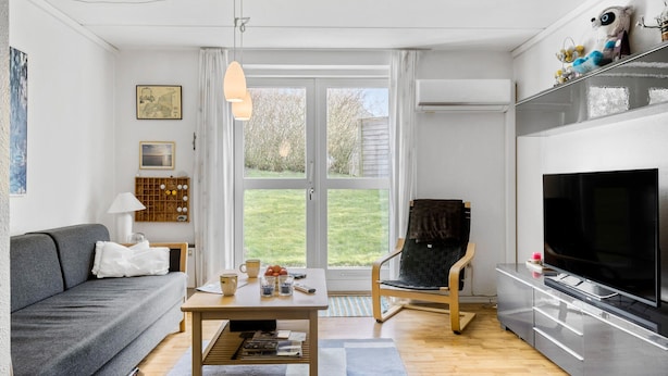Ein modernes Wohnzimmer mit einer Couch, einem Couchtisch, einem Fernseher und Blick auf den Garten durch große Fenster.