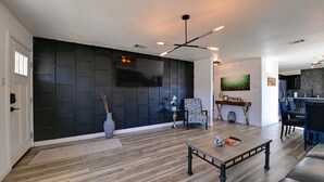A modern living room with a large flat-screen TV, dark paneling on the wall, a dining area with a bar, and a kitchen in the background. - 'Little Elm Diamond' - Steps to Lewisville Lake! (Little Elm)