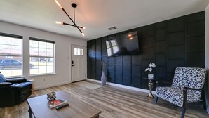A modern living room with a wooden floor, a black accent wall, a mounted TV, a black leather sofa, a patterned armchair, and a coffee table with a book and remote control. - 'Little Elm Diamond' - Steps to Lewisville Lake! (Little Elm)