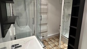 A modern bathroom with a glass shower enclosure, a white bathtub, and a black shelving unit. - Studio Apartment für 2 - Guestrooms24 - Marl (Marl)