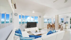 A modern living room with a large TV, white sofas with blue cushions, a coffee table, and a dining area with wooden chairs. - Casa Caribe: NEW beachfront, oceanview condo w/ housekeeper & rooftop terrace (Punta Cana)
