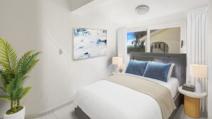 A modern hotel room with a large bed, a potted plant, a painting, and a window with a view of palm trees. - Casa Caribe: NEW beachfront, oceanview condo w/ housekeeper & rooftop terrace (Punta Cana)
