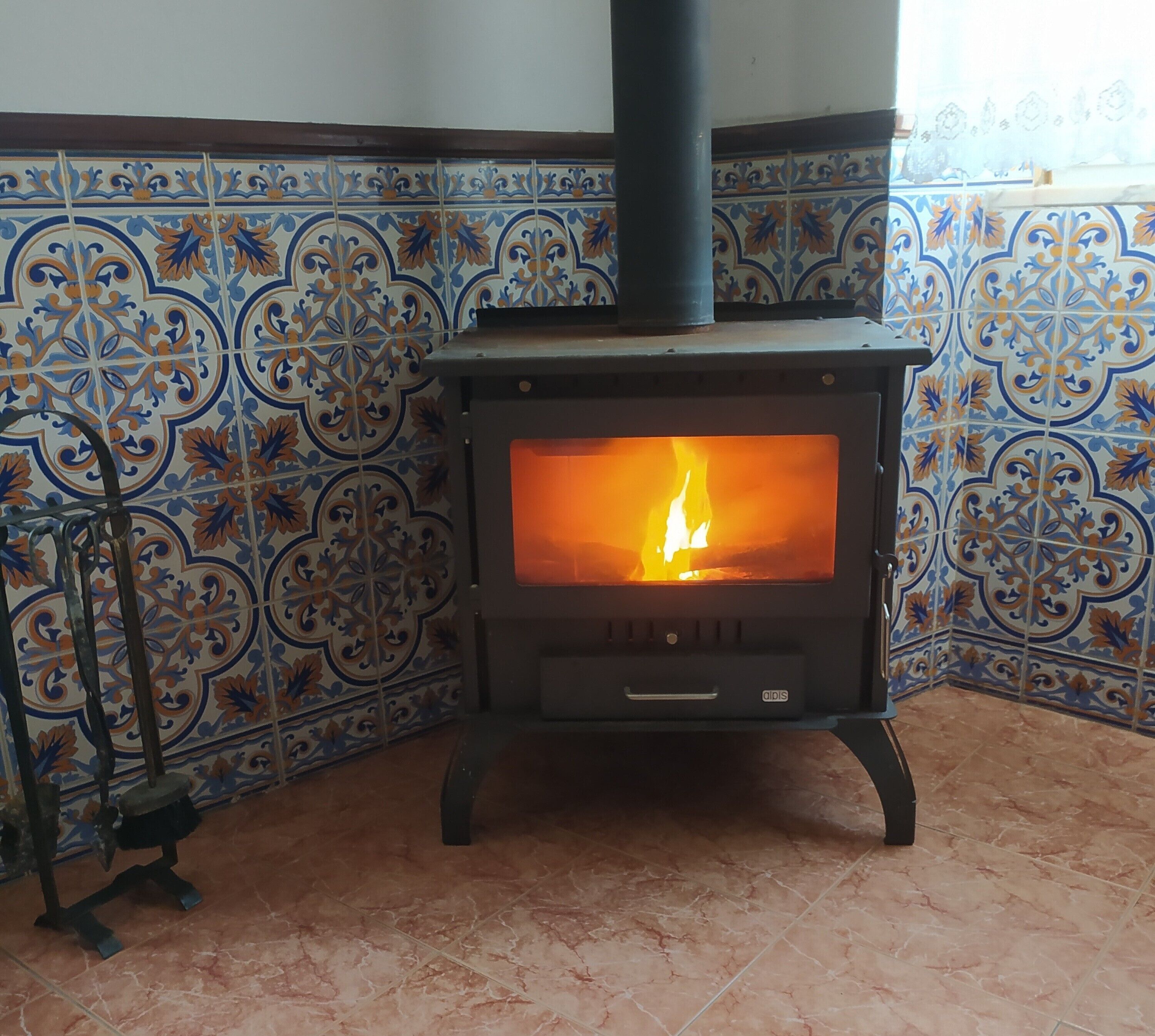 A wood-burning stove with a visible fire, set against a tiled wall with a blue and orange pattern.