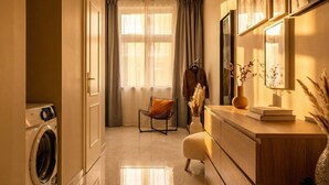 A modern hotel room with a washing machine, a chair, a vanity with a mirror, and a large window with curtains.