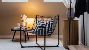 A modern room with a black wireframe chair, a wooden side table, a floor lamp, and a tall vase with white feathers.
