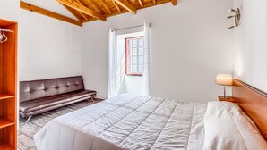 Un dormitorio con techo de madera, una cama con edredón blanco, un banco, un armario y una ventana con cortinas.