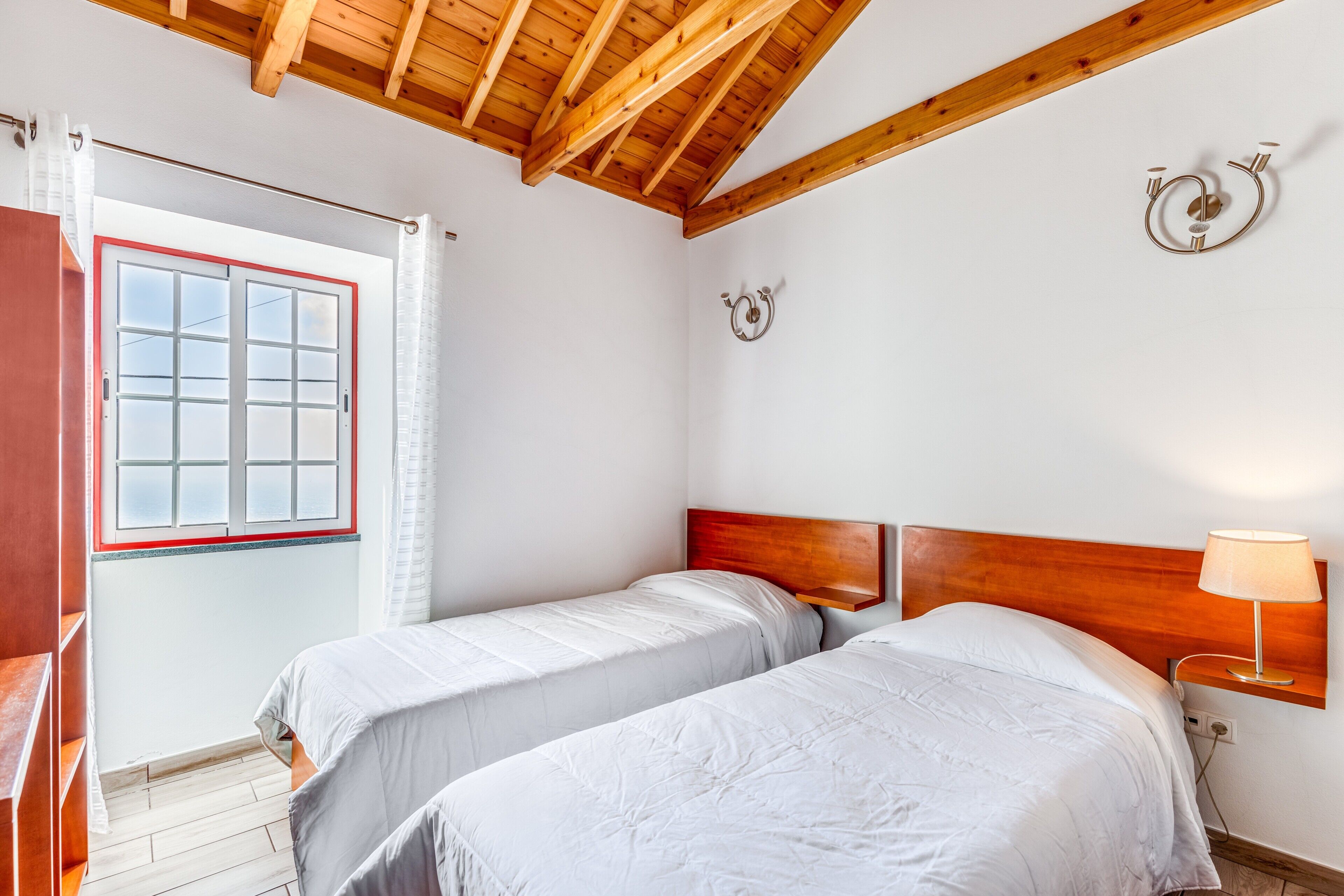 Un dormitorio con dos camas, techo de madera, ventana con cortinas y una lámpara fijada a la pared.