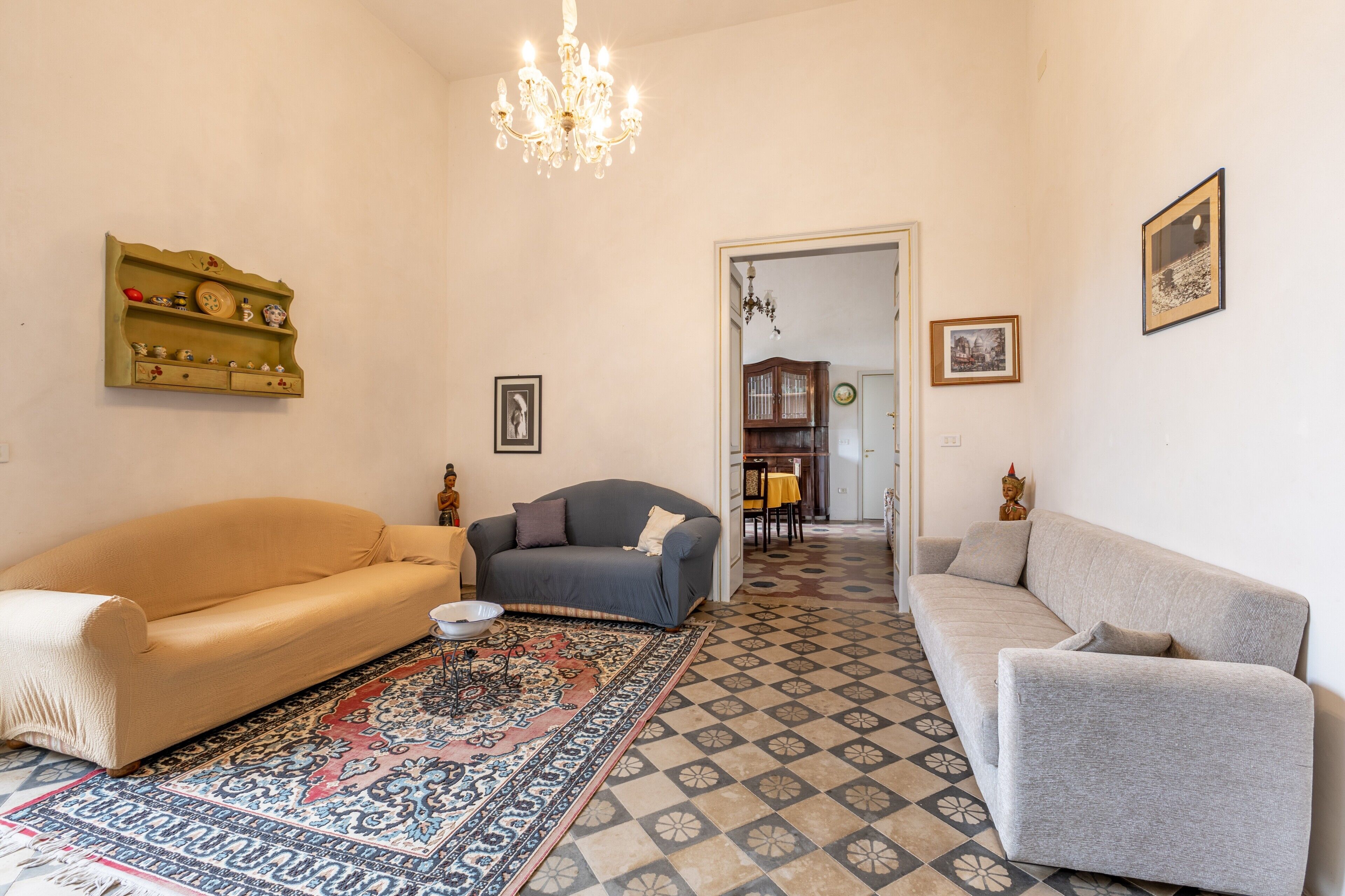 A living room with a beige sofa, a grey sofa, a patterned rug, a chandelier, and a wooden shelf with decorative items.