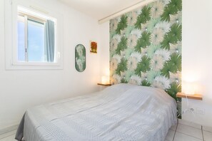 A bedroom with a bed, a window with curtains, a wall-mounted shelf with a lamp, and a wall with a leaf-patterned wallpaper. - Apartment 'Appartement Canet Plage Front Mer' with Sea View, Private Terrace and Air Conditioning (Canet Plage)