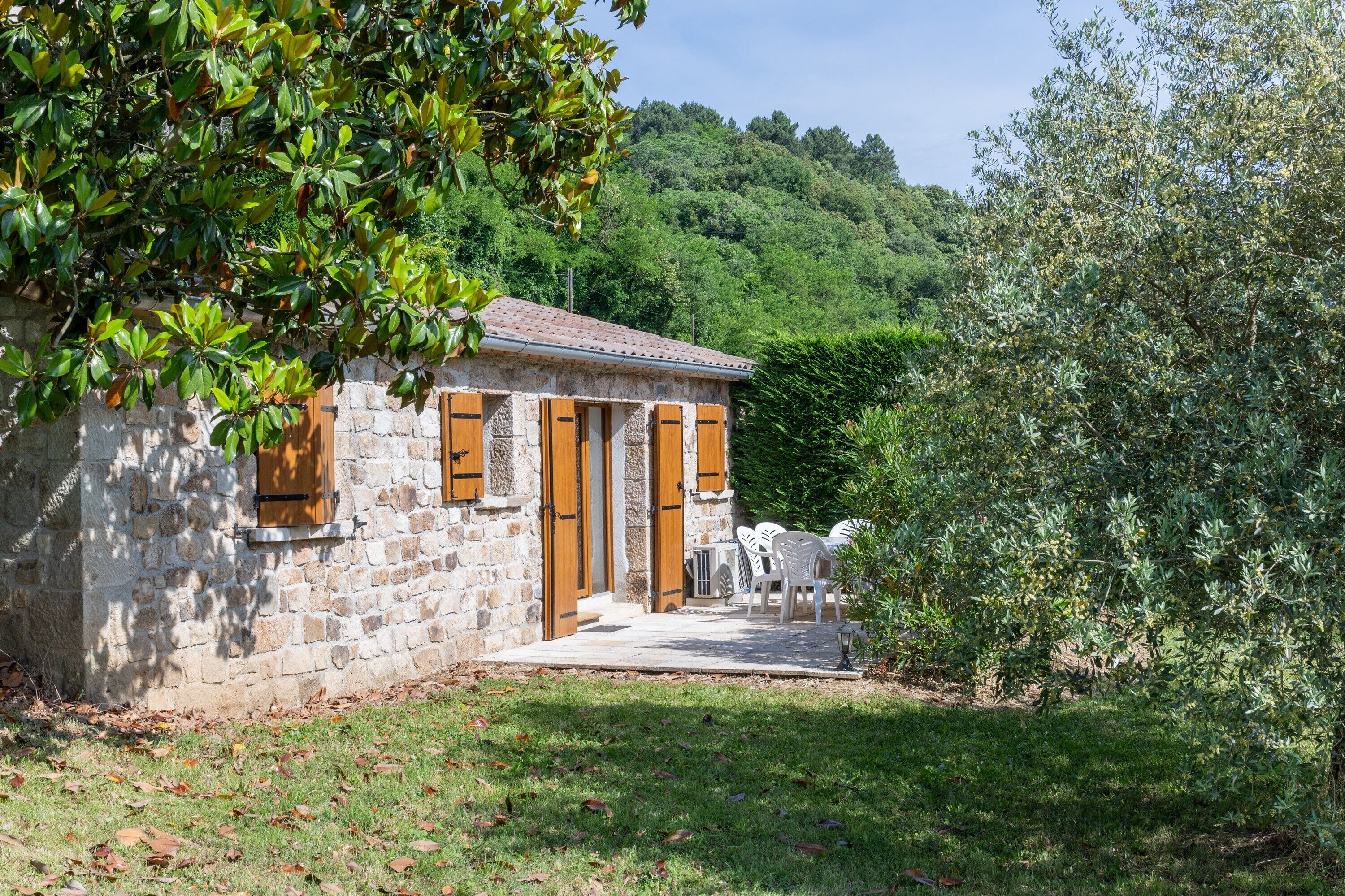 A stone building with a tiled roof, wooden shutters, and a small patio with white plastic chairs.