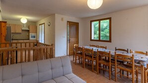 A dining area with a table and chairs, a sofa, a kitchen with wooden cabinets, and a window with a view of greenery. - Les Sources Gite Cabernet House with Shared Pool, Private Terrace, and Air Conditioning (Chambonas)