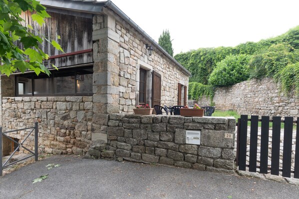 A stone building with a wooden door, a small outdoor seating area with a table and chairs, and a gate. - Charming 'Les Sources Gîte La Grange' House with Shared Pool and Private Terrace (Chambonas)