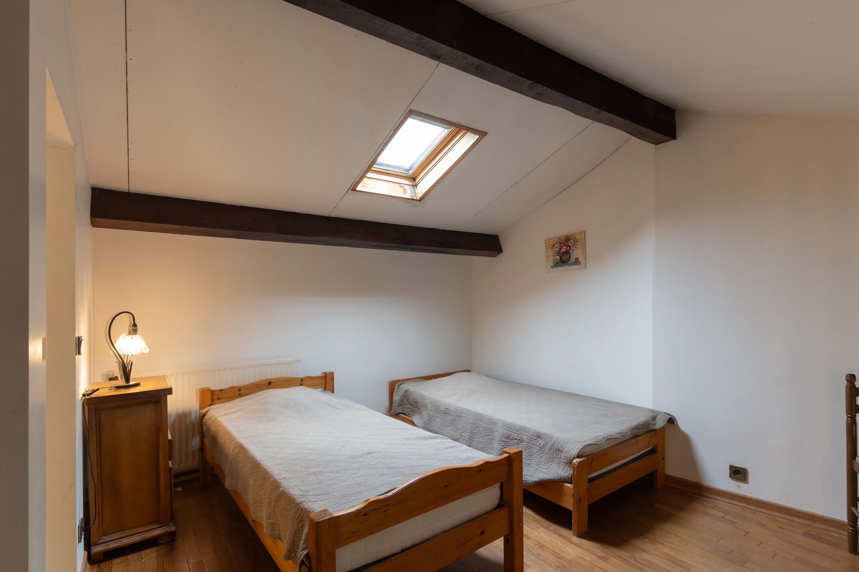 A dormitory room with two wooden beds, a skylight, a bedside table with a lamp, and a radiator.