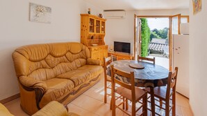 A living room with a sofa, a dining table with chairs, a TV, a wooden cabinet, and a window with a view of a house. - 'Les Sources Gîte La Remise' House with Shared Pool, Private Terrace, and Air Conditioning (Chambonas)