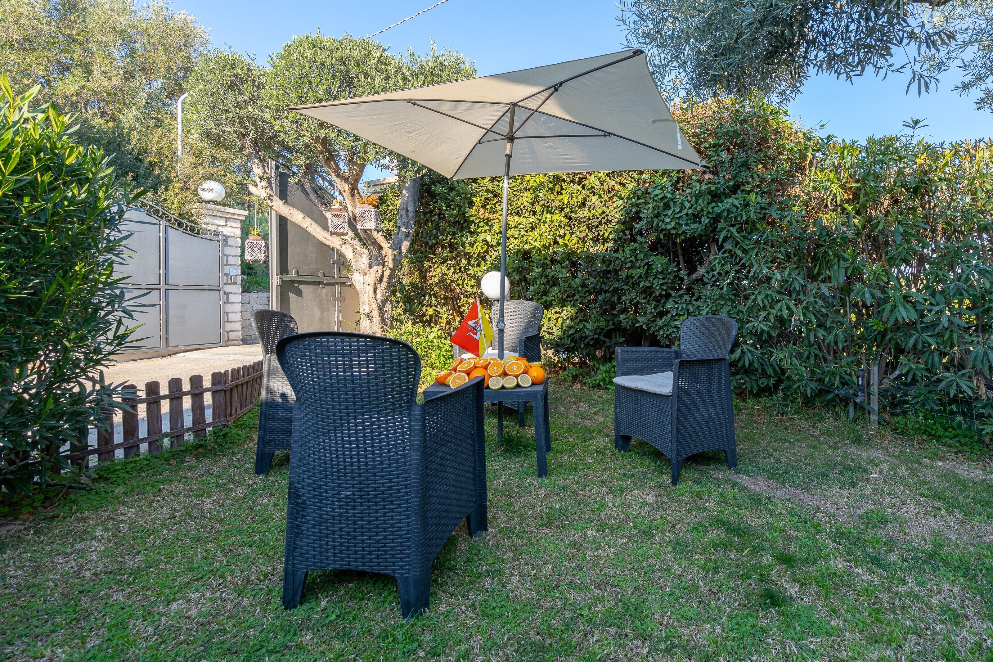 An outdoor seating area with wicker chairs and a large umbrella, surrounded by greenery and a wooden fence.