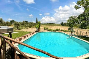 A rectangular swimming pool with a wooden deck and metal railings, surrounded by a grassy area with trees and a clear sky.