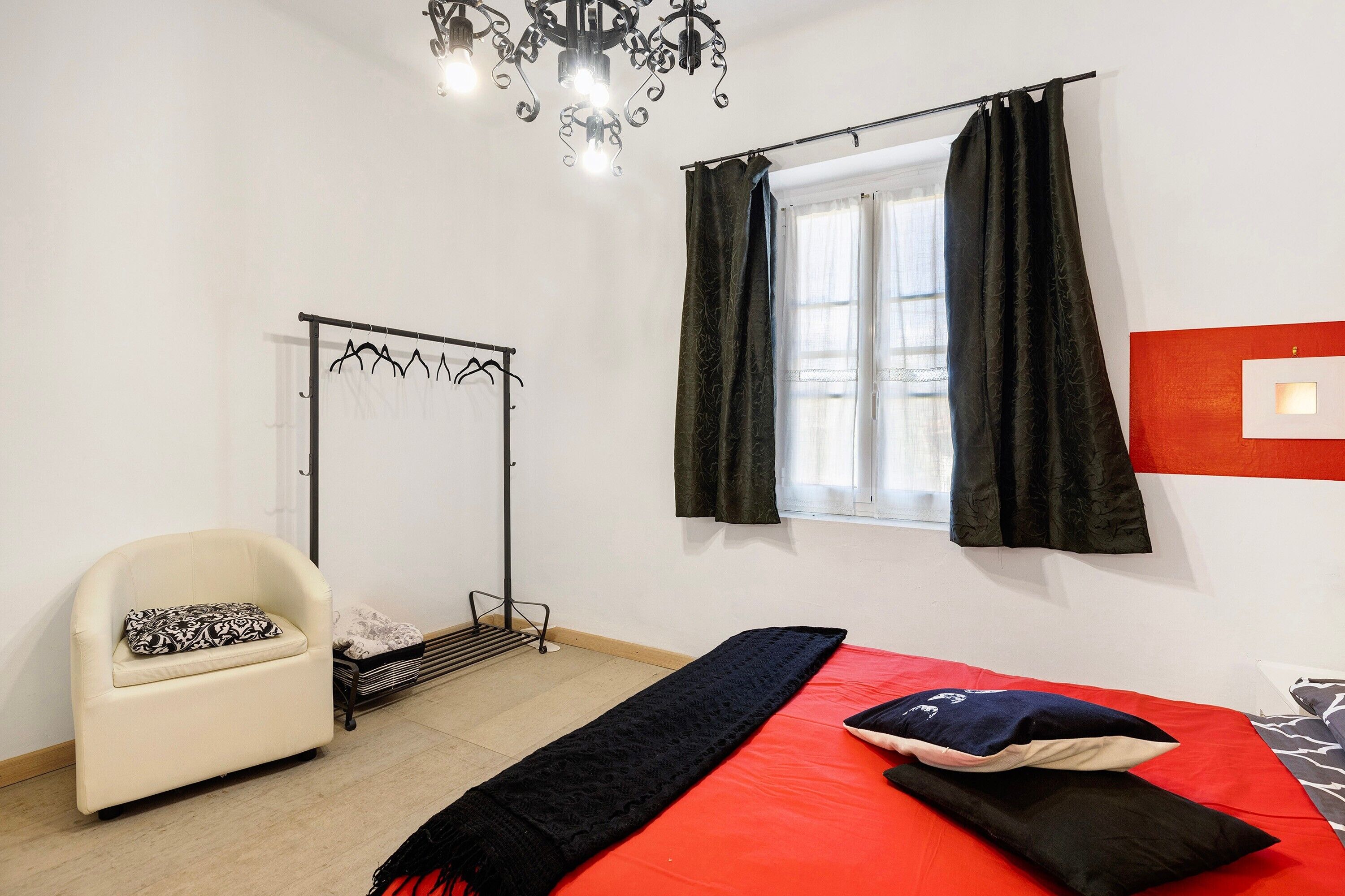 Un dormitorio con una cama, una silla, un radiador, una ventana con cortinas y una araña.
