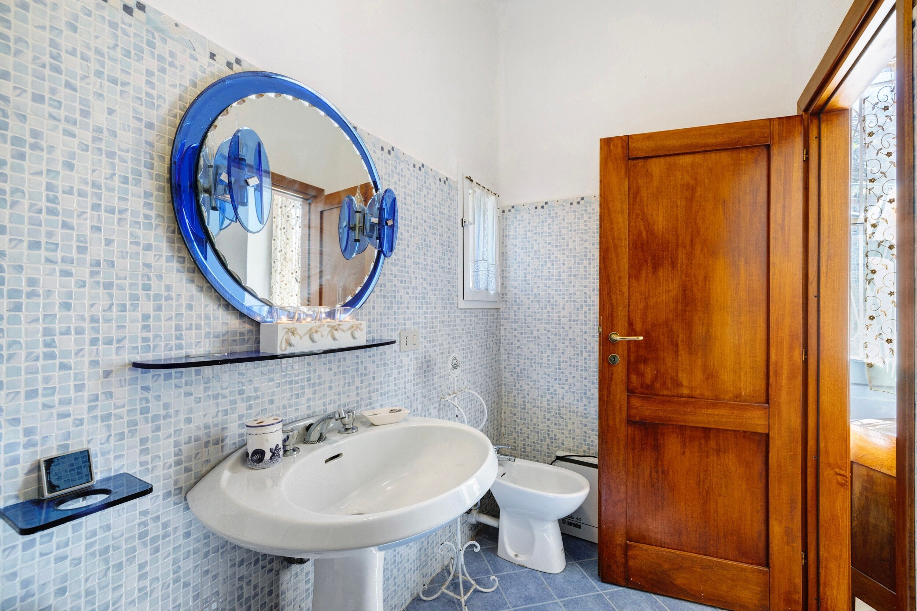 Ein Badezimmer mit einem weißen Waschbecken, einem runden Spiegel, einem Bidet und einer Holztür.