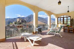 A balcony with sun loungers, a table, and a view of a castle and mountains.