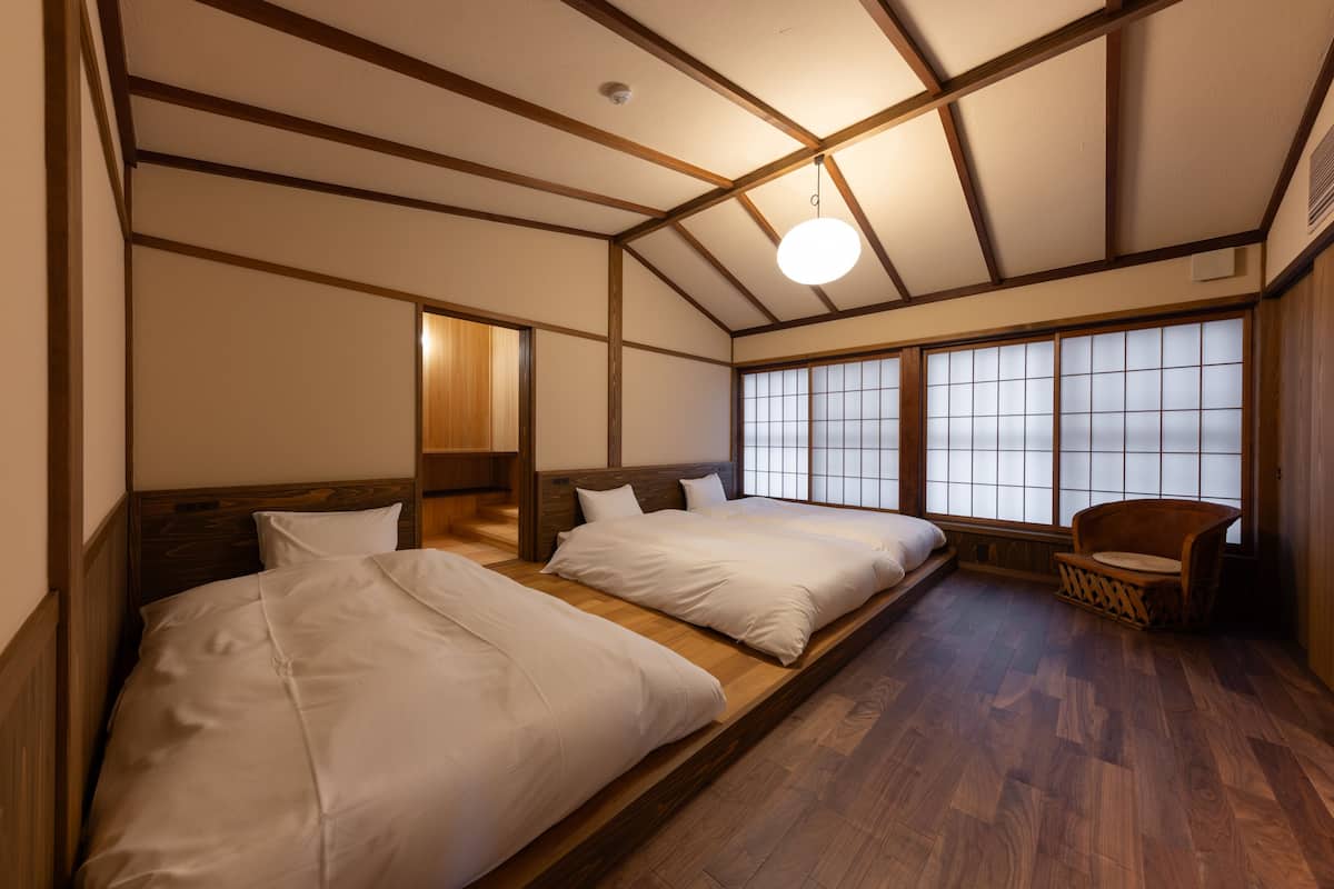 Una habitación tradicional japonesa con suelo de tatami, dos camas con ropa blanca, cabeceros de madera, una ventana con paneles shoji y una pequeña silla de madera.