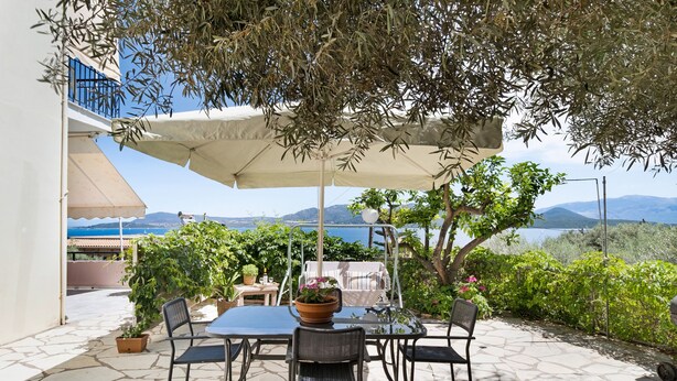 Une terrasse extérieure avec une table et des chaises, un grand parasol, des plantes en pot et une vue sur la mer et les montagnes.