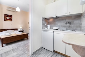 A bedroom with a wooden bed, a bedside table with a lamp, and a small nightstand. Adjacent is a kitchenette with a sink, a stove, and a dishwasher.