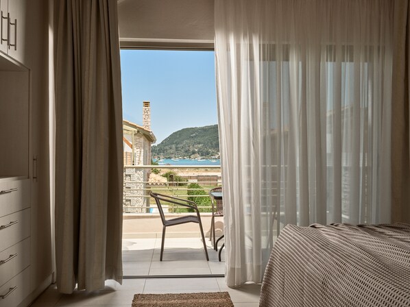 A bedroom with a balcony, a bed, a chair, and a view of a coastal area.