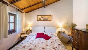 Une chambre avec un plafond en bois, un lit recouvert d’une literie à motifs floraux, deux lampes de chevet, une table de chevet, une commode et une fenêtre avec des rideaux.