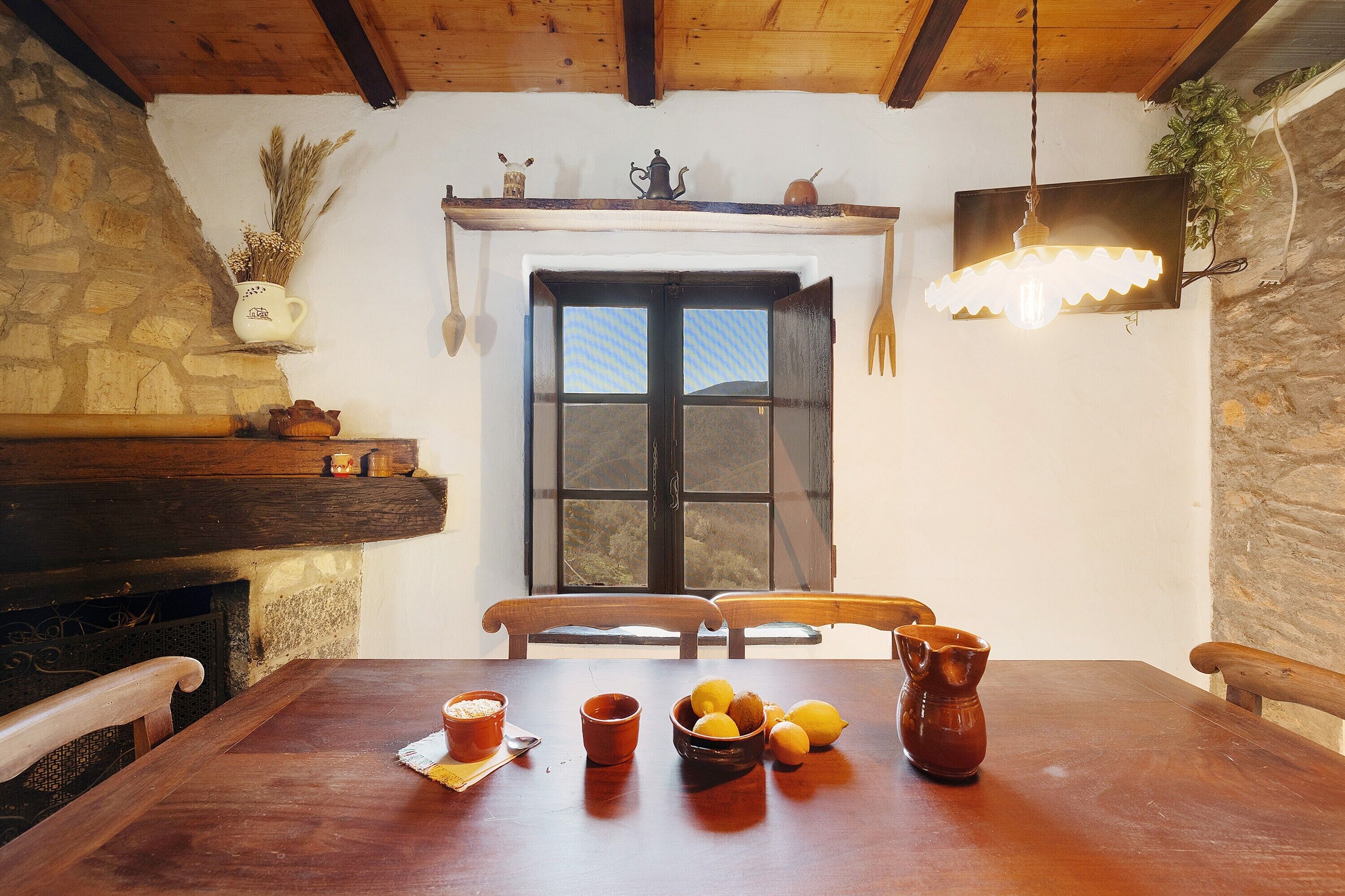 A rustic dining area with a wooden table, chairs, a stone wall, a window, and a hanging light fixture.
