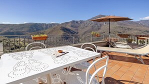 Une terrasse avec une table blanche, des chaises et un parasol, d’où l’on peut admirer un paysage montagneux.