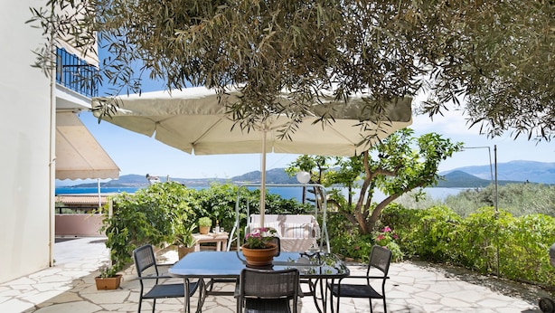 Eine Terrassensitzgruppe im Freien mit Tisch, Stühlen, einem großen Sonnenschirm, Topfpflanzen und Blick auf das Meer und die Berge.