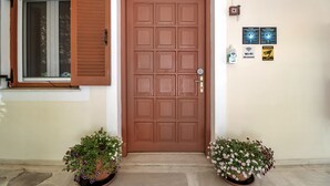 Une porte marron avec une fenêtre au-dessus, deux plantes en pot de chaque côté et un mur sur lequel sont accrochés deux panneaux.