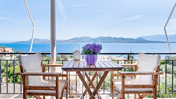 A balcony with two wooden chairs, a small table, a potted plant, and a view of the sea and mountains.