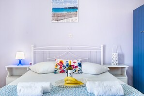 A bedroom with a bed, a nightstand with a lamp, a decorative pillow, a tray with bananas and a bottle, and a wall with a beach-themed poster.