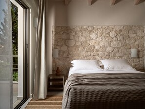 Un dormitorio con pared de piedra, una cama con sábanas blancas, una mesita de noche con un florero y vistas a través de una ventana con cortinas.