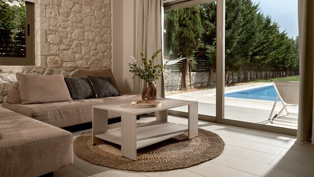 A modern living room with a stone wall, a beige sofa, a coffee table, and a view of a swimming pool. - Villa 'Avesta Private Villas - Kallisto' with Sea View, Private Pool and Air Conditioning (Lefkada)