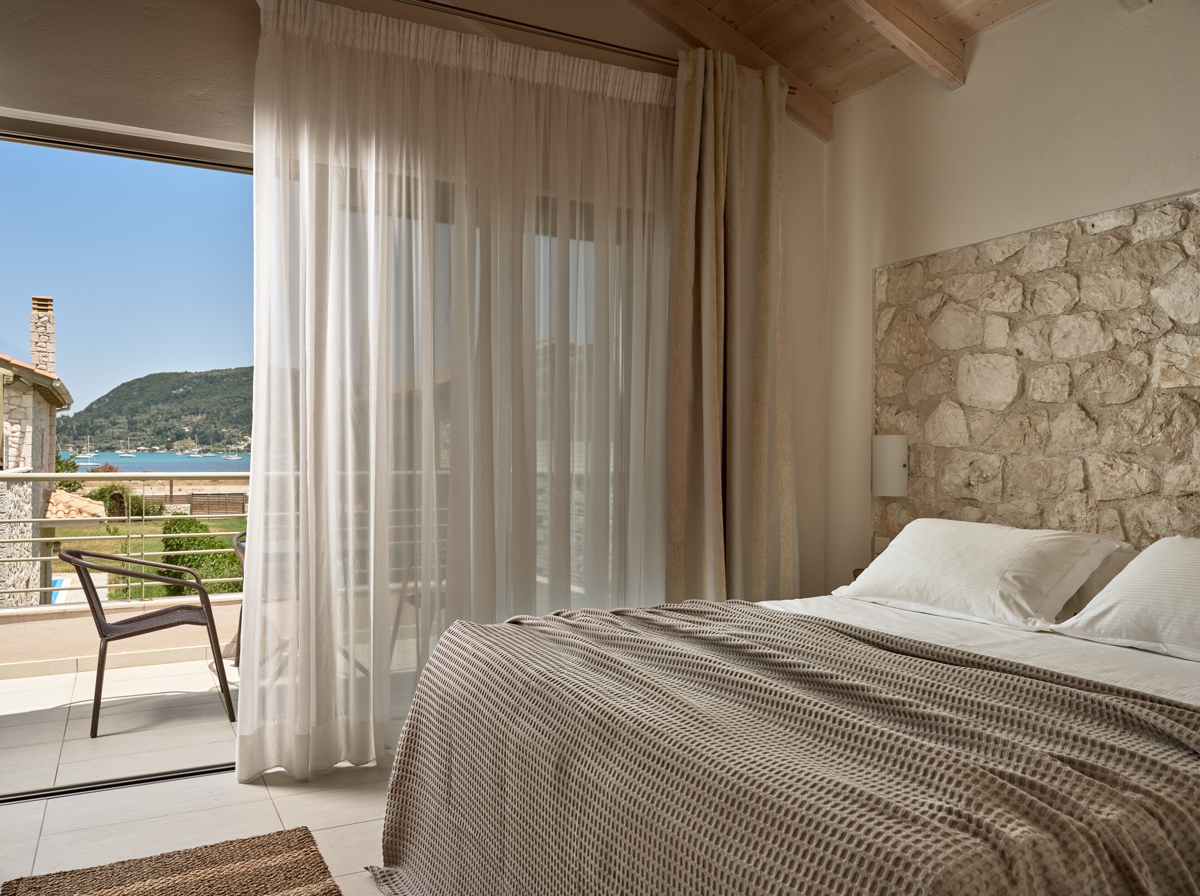 A bedroom with a stone wall, a bed with white linens, a view of the sea, and a balcony with a chair.
