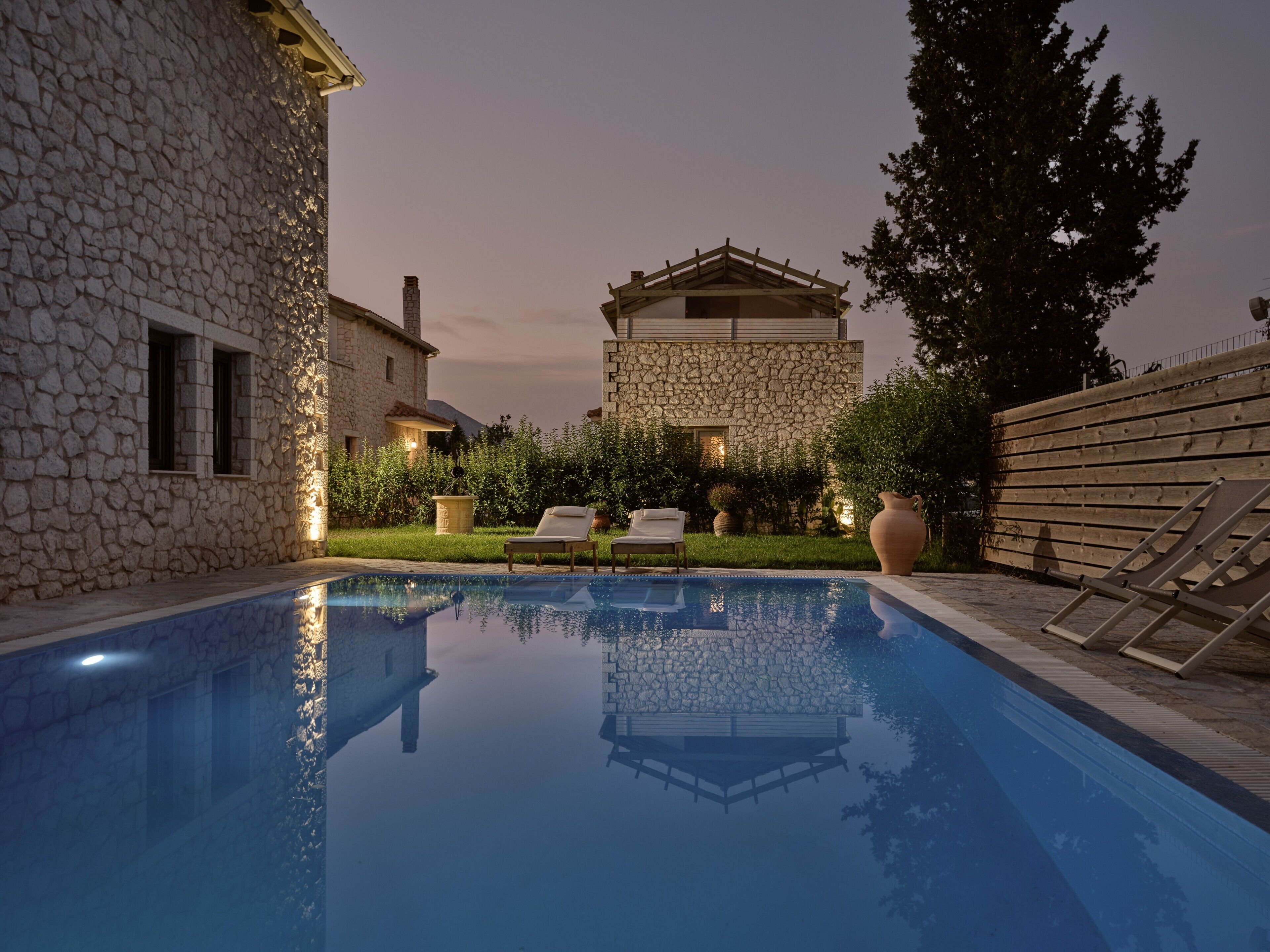 A swimming pool with lounge chairs, stone buildings, and a wooden fence.