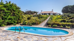 Une piscine aux bords recouverts de pierres, entourée de verdure, et un chemin menant à une maison dont le toit est fait de tuiles rouges.