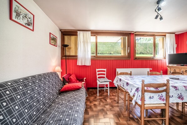 Ein Zimmer mit einer Couch, einem Esstisch mit Stühlen, einem Fernseher und einem Fenster mit Vorhängen.