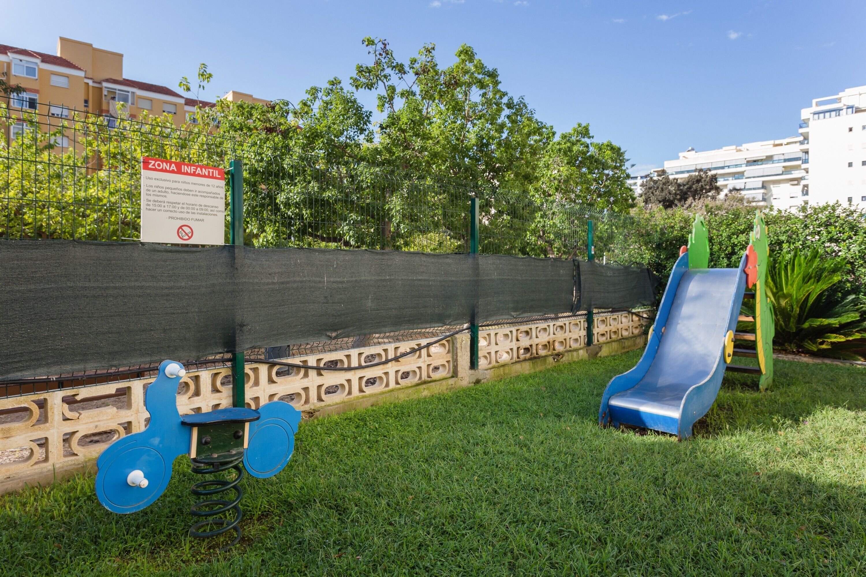 A playground with a slide and a swing set, surrounded by a fence and greenery.