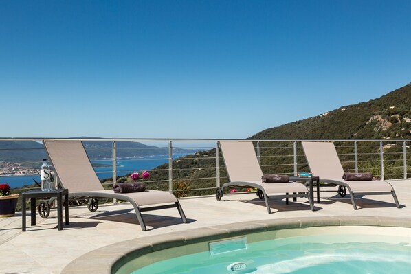 A poolside area with lounge chairs, a table with a bottle, and a view of the sea and hills.