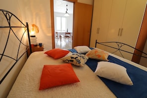 A bed with multiple pillows, a bedside table with a lamp, and a view into a dining area.