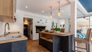 A modern kitchen with a central island, a built-in oven, and a dining area with a wicker chair. - Villa 'L'acanthe' with Private Pool, Private Terrace, and Wi-Fi (Six-Fours-les-Plages)